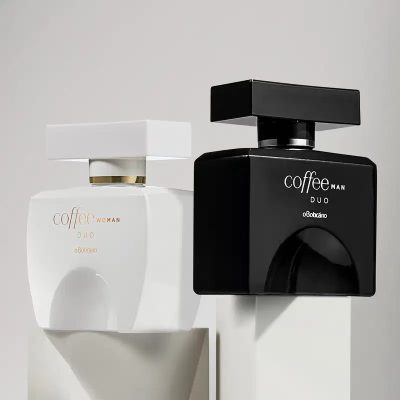 Dois vidros quadrados com tampa retangulares, detalhes e letras douradas, contendo desodorante colônia Coffee Duo, sendo um preto da linha Man e outro branco da linha Woman sobre plataformas brancas em fundo cinza.