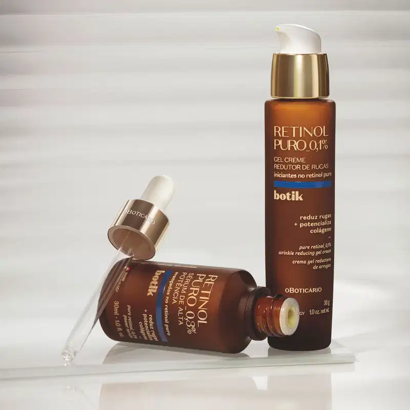 Dois frascos de Retinol Puro 0,1% Botik em embalagem marrom com detalhes dourados e azuis, um com conta-gotas aberto, dispostos sobre superfície branca refletiva com fundo cinzento claro.