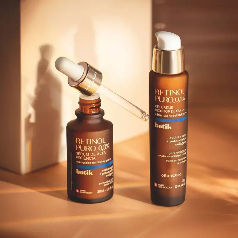 Dois itens Botik Retinol Puro em tons de marrom, dourado, branco, bege e azul, sendo um frasco de gel creme redutor de rugas e um frasco com conta-gotas de sérum facial sobre superfície bege com sombras.