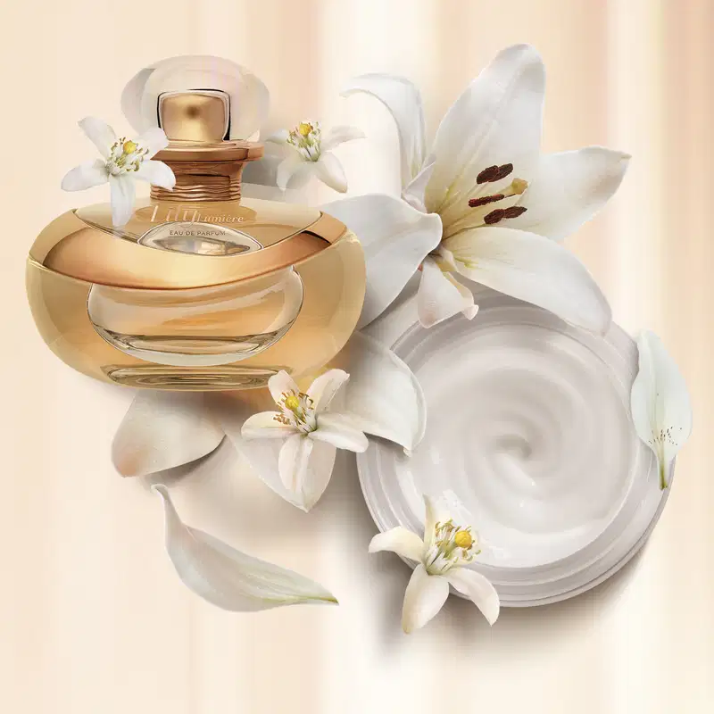 Dois itens Lily Lumière, sendo um vidro arredondado com detalhe dourado, líquido bege, e tampa cristal de Eau de Parfum e pote com creme branco, tampa e letras douradas de creme acetinado corporal sobre fundo listrado de bege e branco com flores brancas.