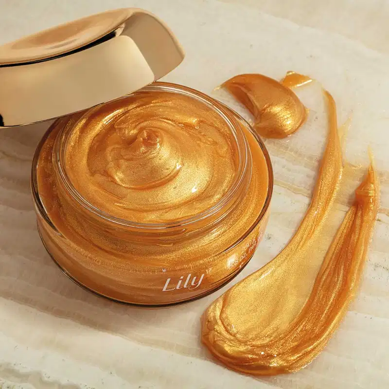 Pote de vidro transparente com creme dourado, tampa dourada e letras bege de geleia iluminadora corporal da marca Lily Glow sobre superfície clara com aplicações do produto espalhadas ao lado e pote branco ao fundo.