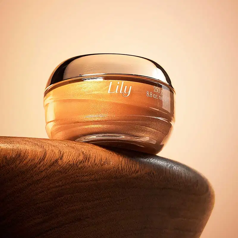 Pote de vidro om creme dourado, tampa dourada e letras bege de geleia iluminadora corporal Lily Glow. posicionado sobre superfície de madeira, com fundo bege claro e iluminação natural que destaca o gel dourado dentro da embalagem com tampa espelhada.