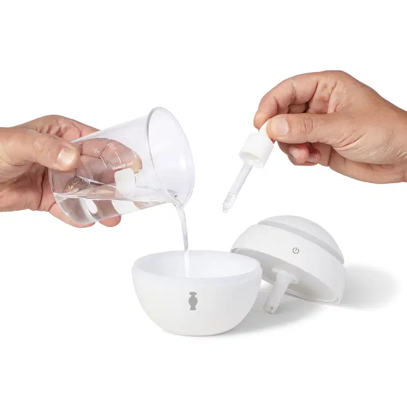 Mão feminina despejando água dentro do pote principal do aromatizador elétrico de ambientes branco. Uma mão masculina pinga essência dentro do mesmo recipiente, enquanto a tampa do aparelho está posicionada ao lado.