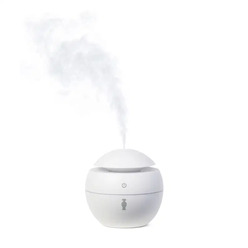 Aromatizador elétrico de ambientes branco com tampa em formato de cúpula, botão power cinza no topo e ícone da marca O Boticário no corpo do pote. Há fumaça branca saindo do topo do aparelho.