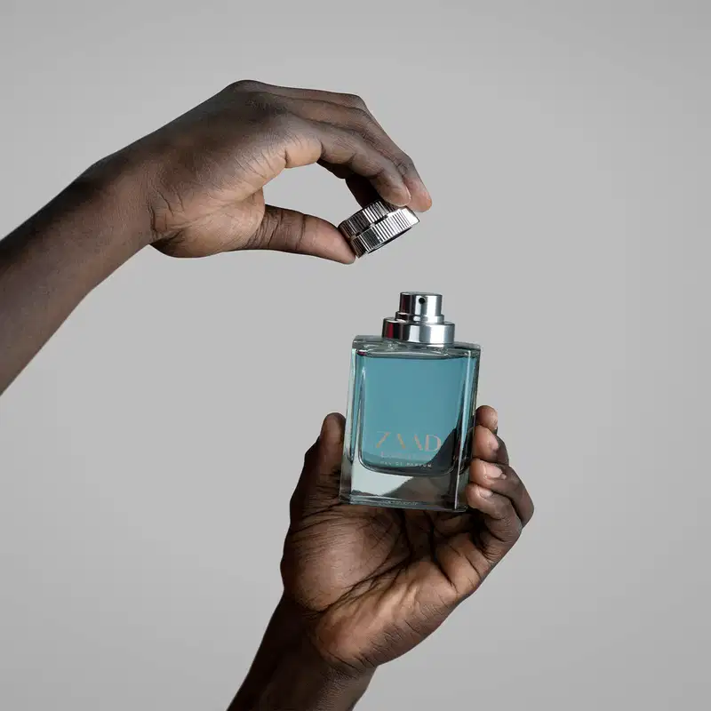 Uma mão masculina segura um Vidro transparente em formato quadrado, com líquido azul acinzentado de Eau de Parfum Zaad Expedition e a outra mão segura a tampa pratada, sobre fundo cinza claro.