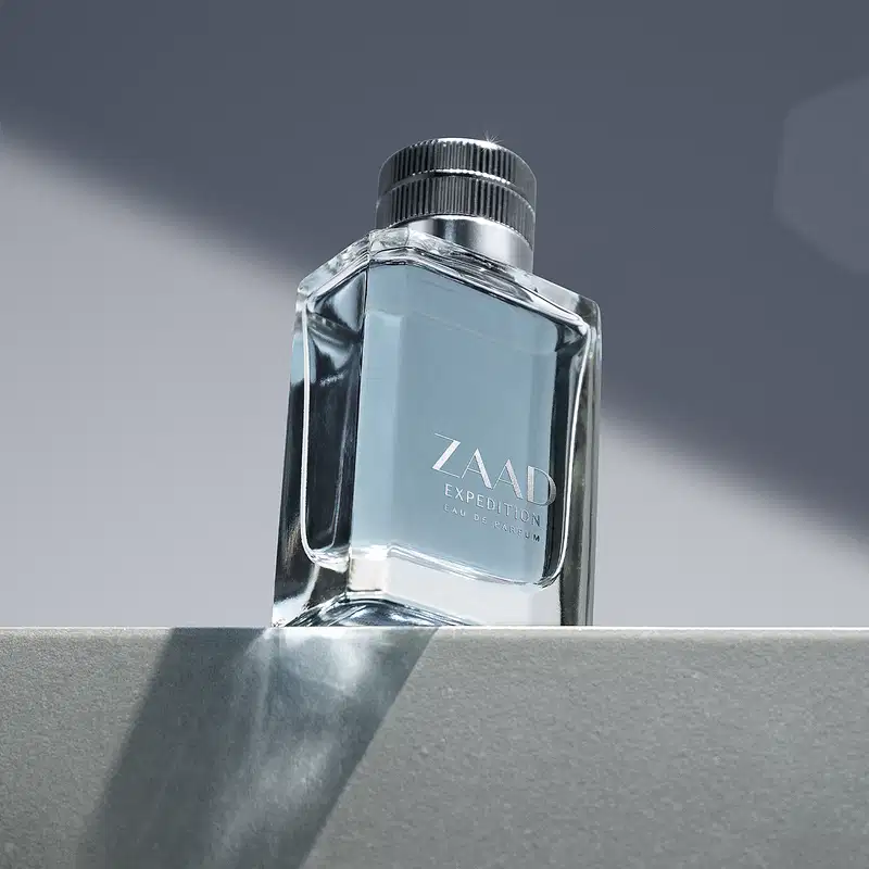 Vidro transparente em formato quadrado, com líquido azul acinzentado, tampa prateada e letras cinzas de Eau de Parfum Zaad Expedition posicionado sobre superfície cinza em ambiente com iluminação natural e sombras.