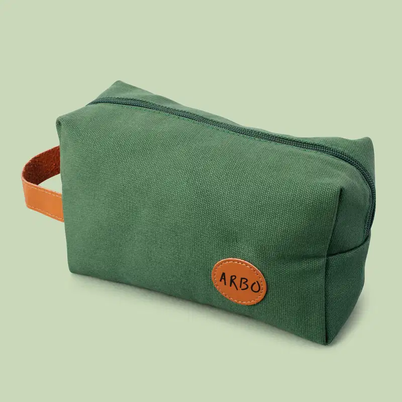 Nécessaire retangular de lona verde com alça de couro caramelo, zíper verde e logo Arbo em patch circular também em couro caramelo sobre fundo cinza.