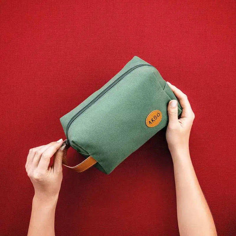 Mãos femininas segurando um nécessaire retangular de lona verde com alça de couro caramelo, zíper verde e logo Arbo em patch circular também em couro caramelo sofre fundo vermelho escuro.