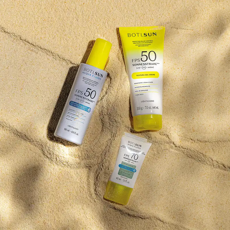Três produtos Boti Sun com protetor solar FPS 50 e 70 posicionados sobre areia clara com sombras, incluindo frasco spray, tubo gel-creme amarelo e embalagem compacta.