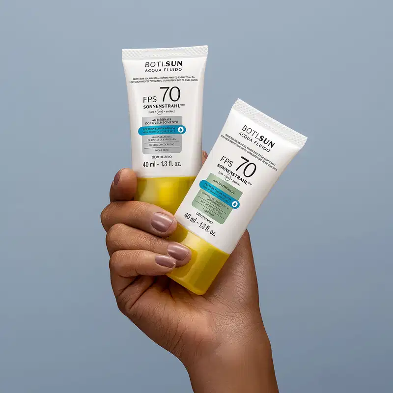 Uma mão feminina segura dois frascos brancos com tampas amarelas, letras pretas e detalhes prata e azul, de protetor solar facial antioleosidade FPS 70 Acqua Fluído da marca Boti.Sun, sobre fundo azul.