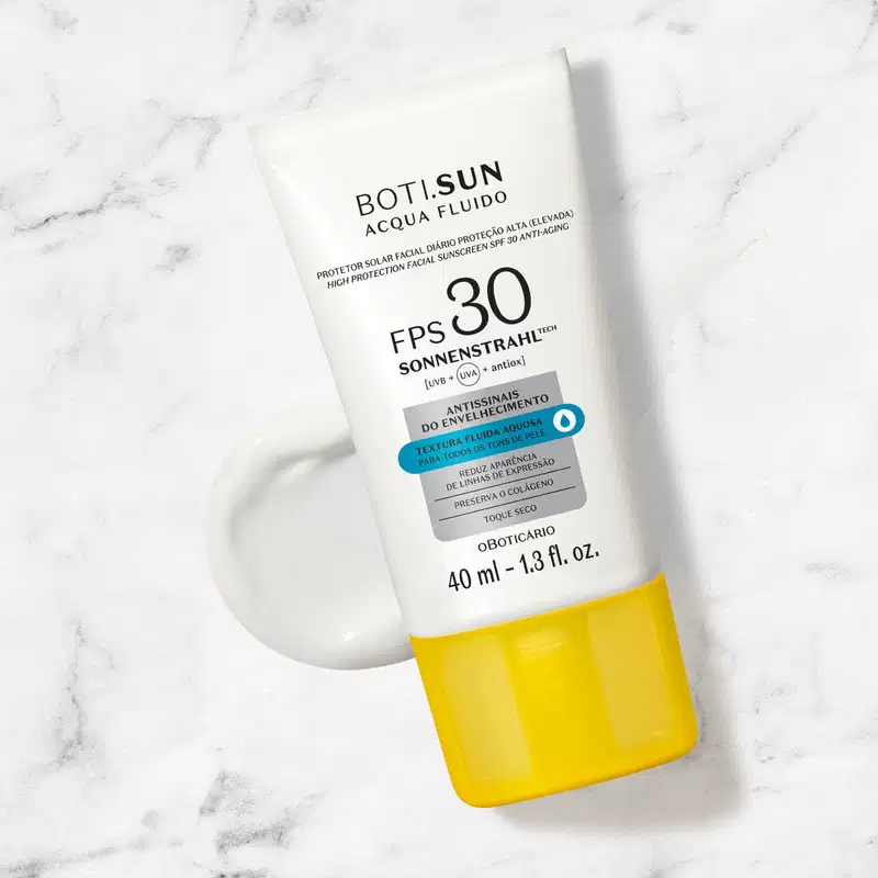Frasco branco com tampa amarela, letras pretas e detalhes prata e azul, de protetor solar facial antissinais FPS 30 Acqua Fluído da marca Boti.Sun posicionado sobre superfície de mármore cinza e branco com sombra projetada ao lado.