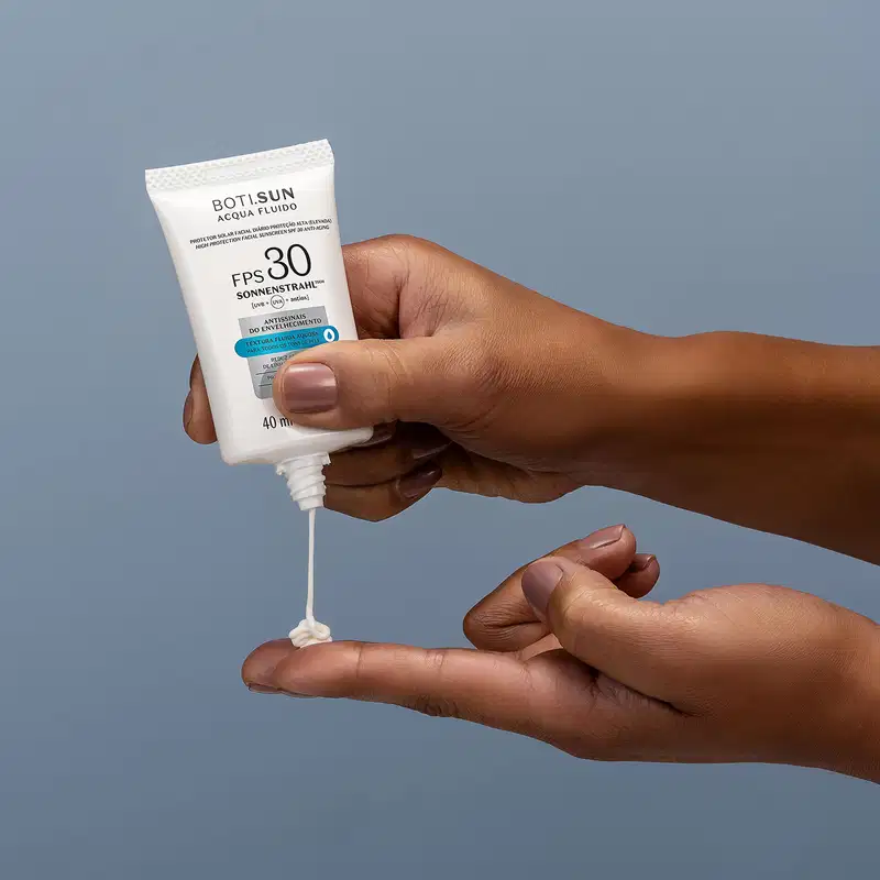 Uma mão feminina segura um frasco branco com letras pretas e detalhes prata e azul de protetor solar facial antissinais FPS 30 Acqua Fluído da marca Boti.Sun e aplica o produto na outra mão, tudo em fundo azul escuro.
