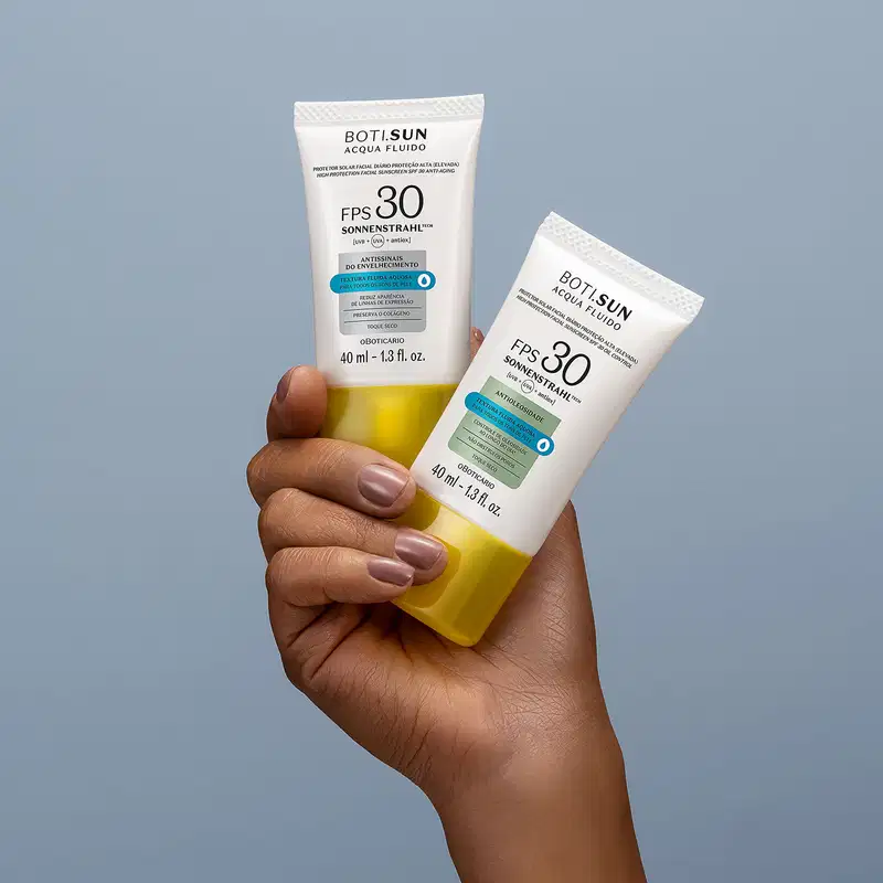Uma mão feminina segura dois frascos brancos com tampas amarelas, letras pretas e detalhes prata e azul, de protetor solar facial antissinais FPS 30 Acqua Fluído da marca Boti.Sun, sobre fundo azul.