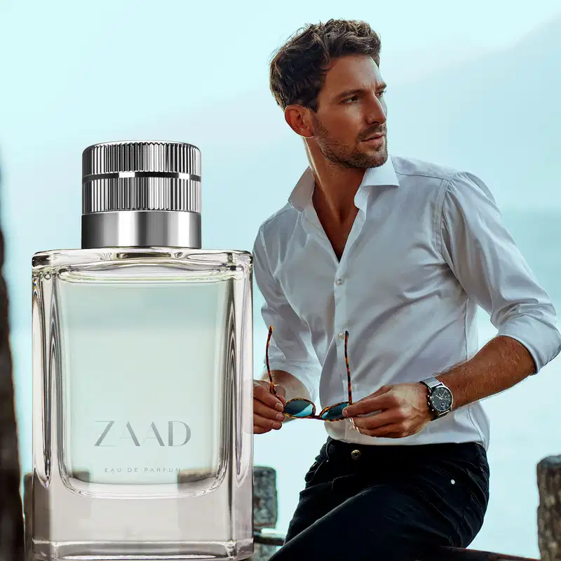 Vidro transparente com líquido cinza, tampa prateada e letras cinzas de Eau de Parfum Zaad ao lado da imagem de um homem segurando seus óculos escuros, usando camisa branca e calça preta, olhando para o lado.