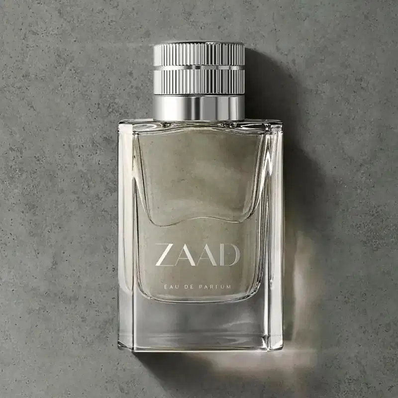 Vidro transparente com líquido cinza, tampa prateada e letras cinzas de Eau de Parfum Zaad, posicionado sobre superfície cinza de concreto com sombra projetada ao lado.