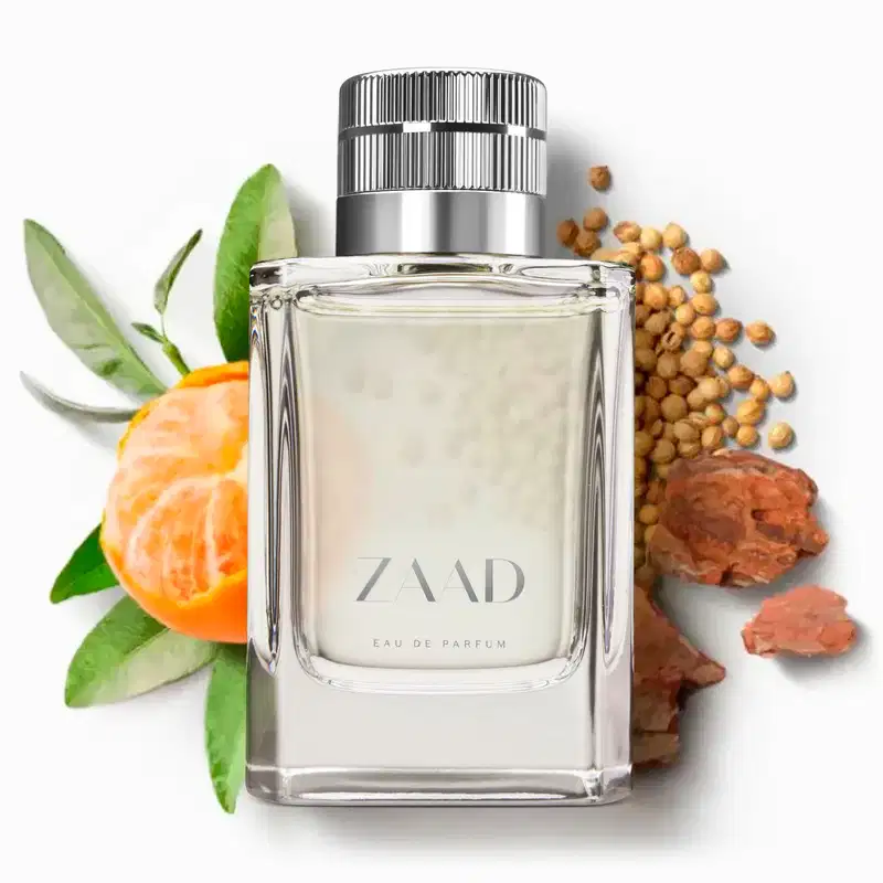 Vidro transparente com líquido cinza, tampa prateada e letras cinzas de Eau de Parfum Zaad sobre superfície branca, cercado por folhas verdes, tangerina, cevada e sementes marrom-avermelhadas.
