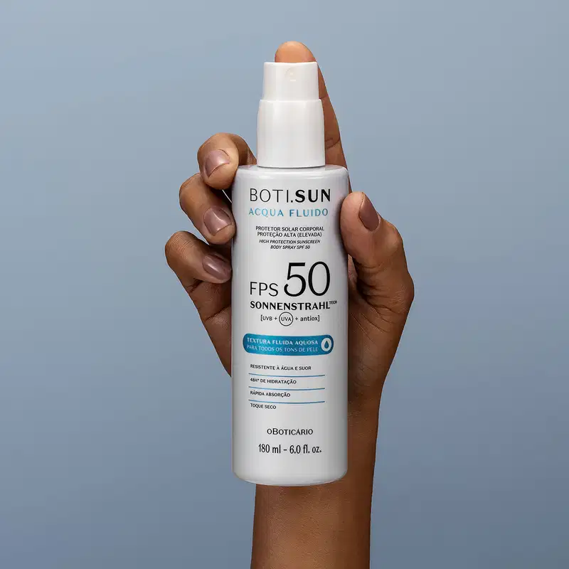 Mão segurando um Frasco branco com tampa amarela, letras pretas e detalhe em azul, contendo 180ml de spray protetor solar corporal FPS 50 Acqua Fluído da marca Boti.Sun em fundo azul petróleo.