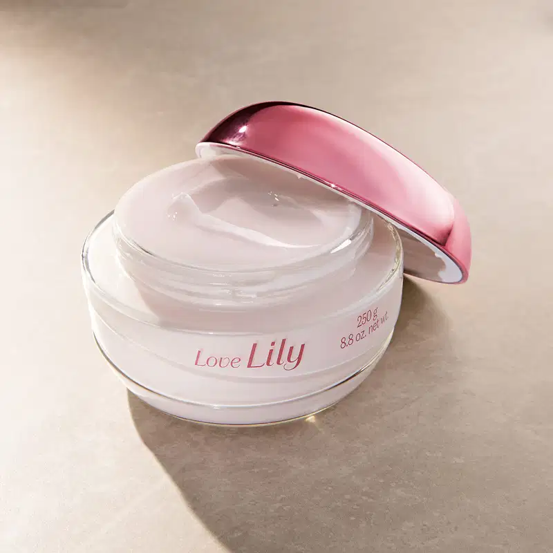 Pote de vidro transparente aberto com creme acetinado hidratante corporal da marca Love Lily na cor branca, tampa roxa encostada ao lado, em fundo lilás com sombra.