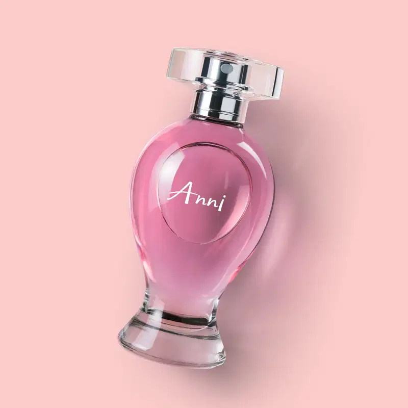 Vidro curvilíneo transparente com liquido rosa, pulverizador prateado e tampa cristal contendo desodorante colônia Boticollection Anni deitado em um fundo rosa.