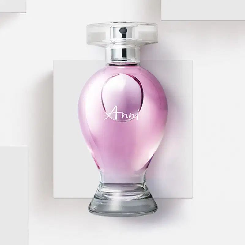 Vidro curvilíneo transparente com liquido rosa, pulverizador prateado e tampa cristal contendo desodorante colônia Boticollection Anni, posicionado sobre superfície branca com painel geométrico ao fundo em ambiente minimalista.