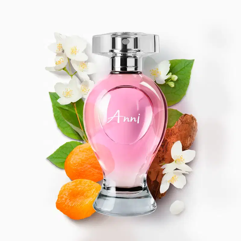 Vidro curvilíneo transparente com liquido rosa, pulverizador prateado e tampa cristal contendo desodorante colônia Boticollection Anni., circundado por flores brancas, frutas cítricas laranja e madeira em fundo branco.