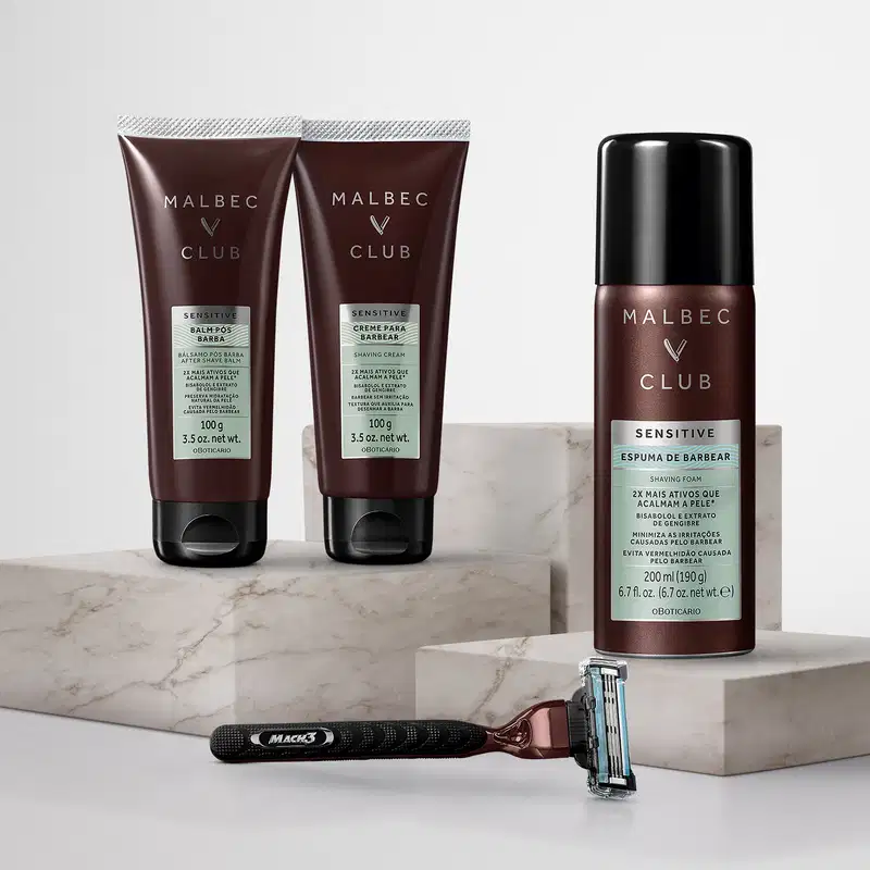 Dois tubos de creme para barbear da marca Malbec Club, em embalagem marrom escura com texto branco. Também há um frasco de espuma de barbear da mesma marca, em embalagem verde clara. Junto, um aparelho de barbear preto com cabo.