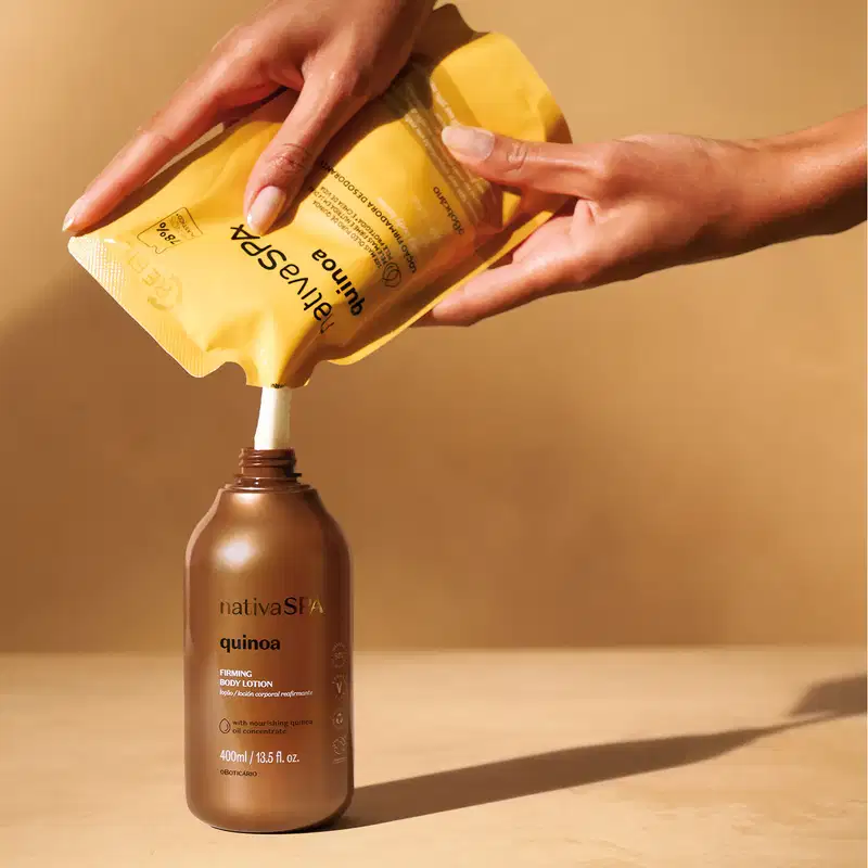 Mãos despejando loção corporal Nativa SPA Quinoa de sachê bege com letras brancas e pretas em frasco dourado sobre superfície bege com sombra natural.