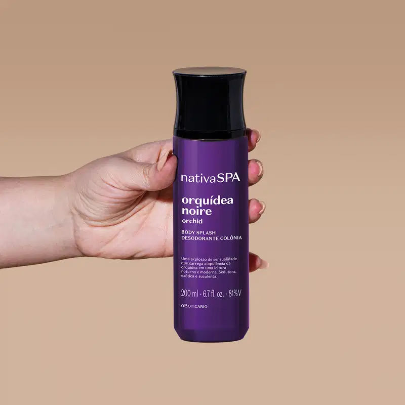 Mão segurando um frasco roxo escuro com tampa preta e letras brancas de body splash da marca Nativa SPA Orquídea Noire em um fundo com tons de bege.