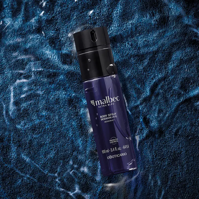 Frasco azul escuro com letras cinzas contendo 100ml de body spray Malbec Ultra Bleu, posicionado sobre superfície azul com textura de fios e padrões geométricos, com partículas brancas dispersas ao redor.