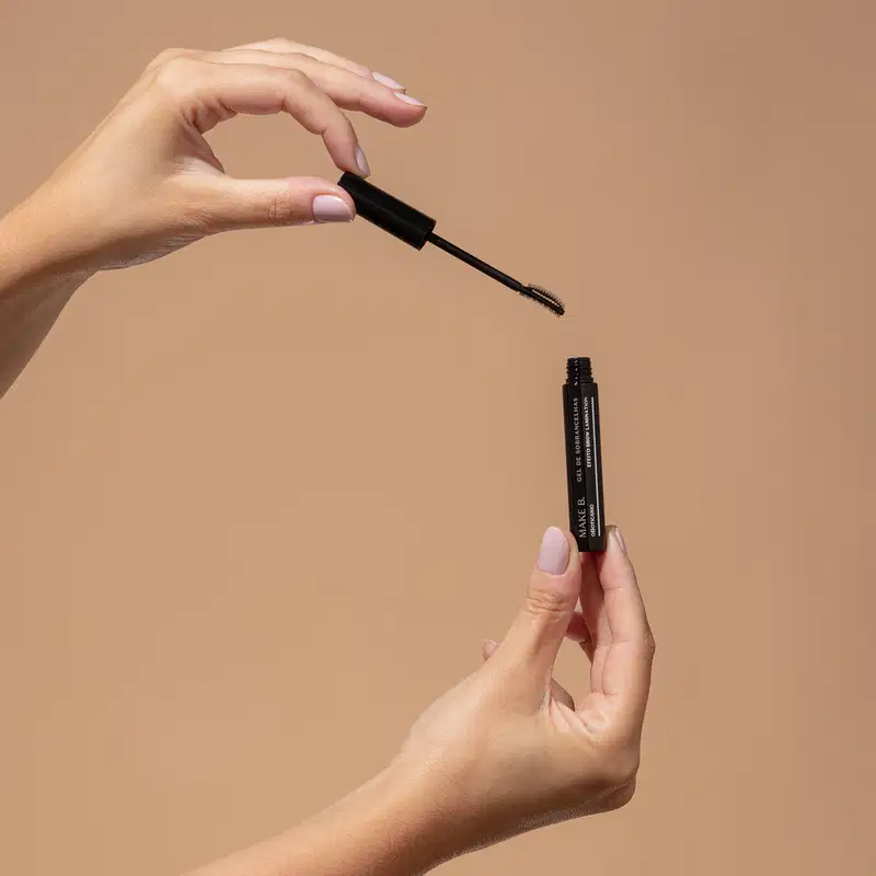 Uma mão feminina segura um tubo preto de máscara de sobrancelhas em gel Make.B Brow Lamination e a outra mão segura a tampa com a haste e a escova aplicadora pretas, sobre fundo em tons de bege.