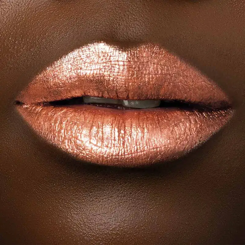 Close-up de um rosto feminino de pele negra evidenciando os lábios dourados e brilhantes, indicando o uso do batom líquido Diamond Dourado Luxury da marca Make B.