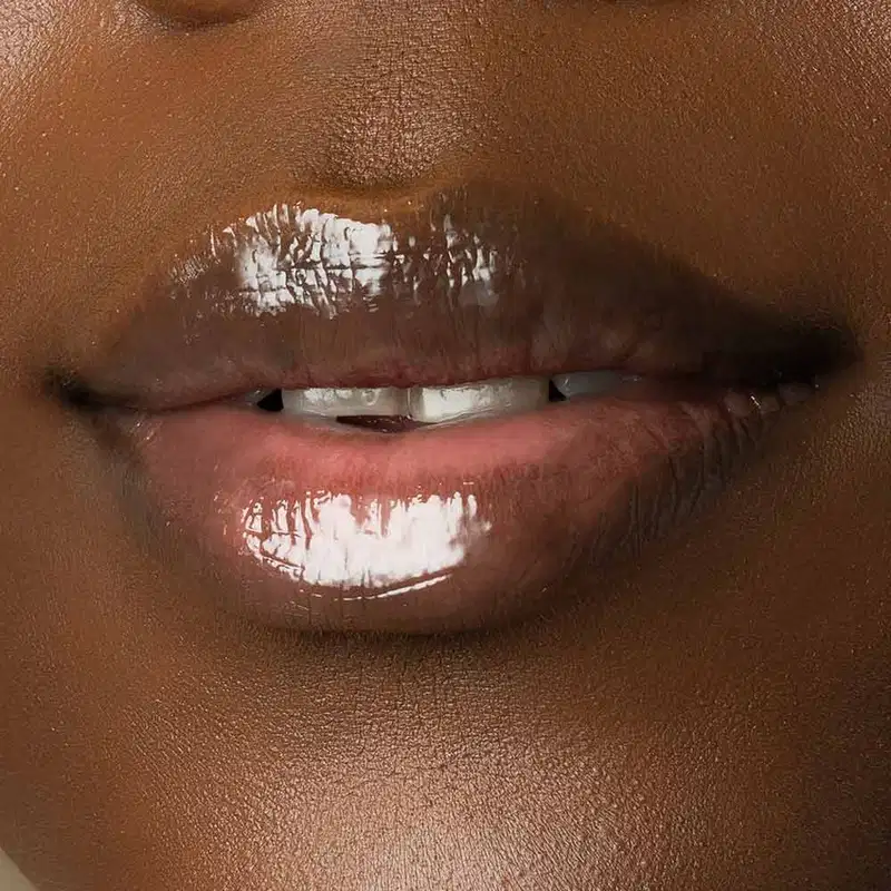 Close-up de um rosto feminino de pele negra evidenciando os lábios brilhantes que usam o gloss labial Incolor Crystal Icon da marca Make B. 