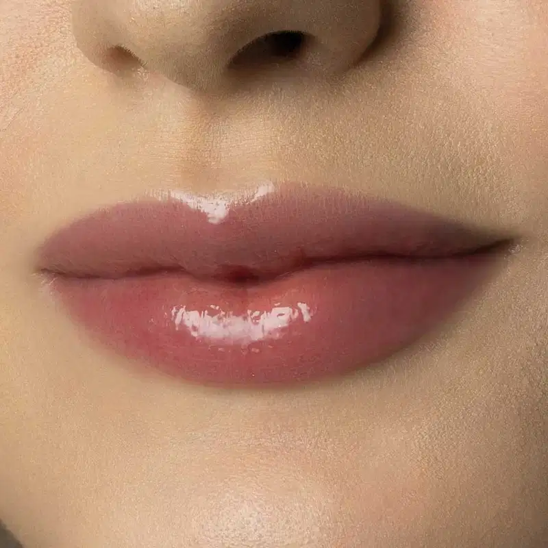 Close-up de um rosto feminino de pele branca evidenciando os lábios brilhantes que usam o gloss labial Incolor Crystal Icon da marca Make B. 