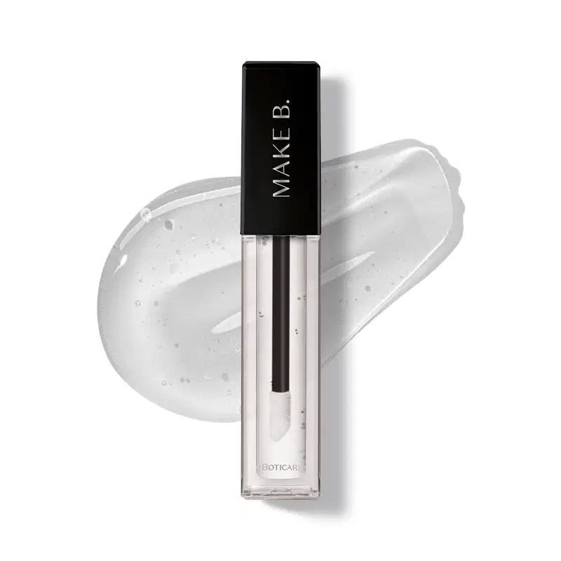 Gloss labial Incolor Crystal Icon da marca Make B. em frasco retangular transparente com líquido brilhante e sem cor e tampa preta com logo da marca, exibido sobre superfície branca com aplicação do produto ao fundo mostrando textura e acabamento.