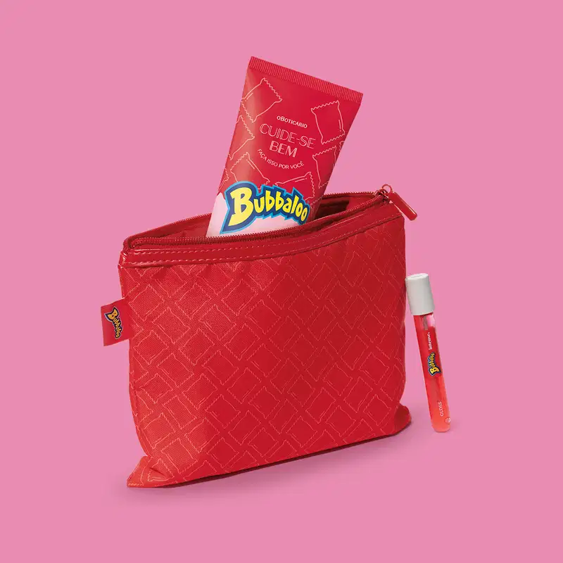 Nécessaire vermelha retangular da marca Cuide-se Bem Bubbaloo com padrão geométrico em dourado, zíper superior e etiqueta da marca Bubbaloo na lateral com um produto de mesma marca dentro, sobre fundo rosa.
