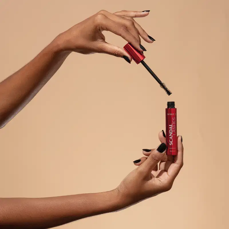 Uma mão feminina segura um tubo vermelho metalizado com letras azuis de máscara de cílios Make B. Scandal Lashes e a outra mão segura a tampa vermelha metalizada com haste e aplicador pretos, sobre fundo em tons de bege.
