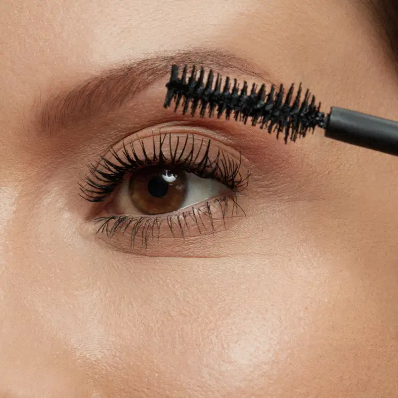 Close-up de um rosto feminino evidenciando os olhos, onde uma escova aplicadora aplica a máscara de cílios Make B. Scandal Lashes nos cílios, deixando os fios alongados e bem definidos.
