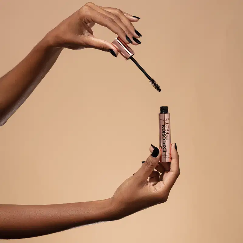 Uma mão feminina segura um tubo rosa furta-cor com letras pretas de máscara de cílios Make B. Explosion Effect e a outra mão segura a tampa rosa furta-cor com haste e aplicador pretos, sobre fundo em tons de bege.