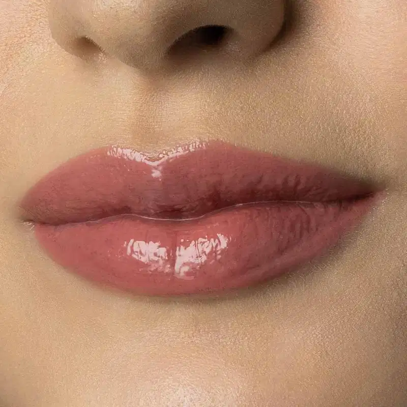 Close-up de um rosto feminino de pele branca evidenciando os lábios brilhantes e rosados que usam o gloss labial Rosa New Rose da marca Make B. 