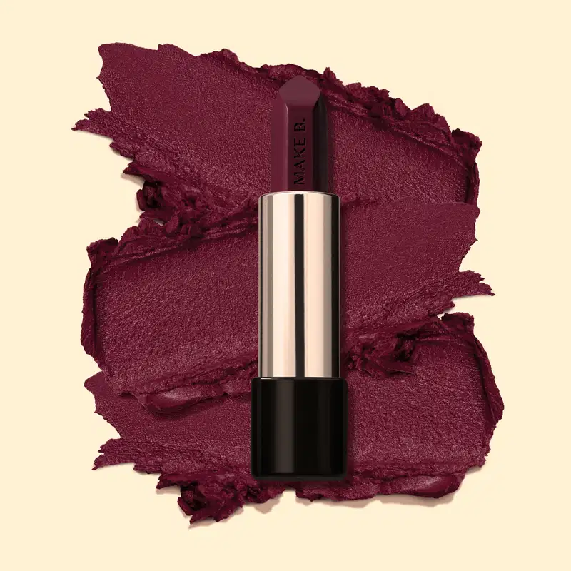 Batom cremoso na cor Vinho Fashion Berry da marga Make B., em tubo prateado com a parte inferior preta posicionado sobre amostra cremosa do produto na cor vinho intenso, sobre fundo bege claro.