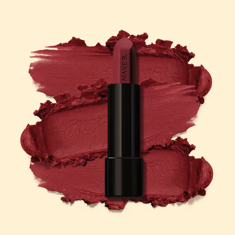 Batom mate na cor Vermelho Red Supreme da marga Make B., em tubo preto posicionado sobre uma amostra do produto na cor vermelho intenso, sobre fundo bege claro.