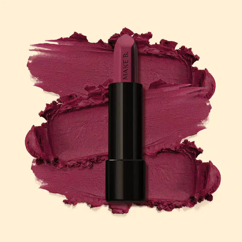 Batom mate na cor Violeta Violet da marga Make B., em tubo preto posicionado sobre uma amostra do produto na cor roxo intenso, sobre fundo bege claro.