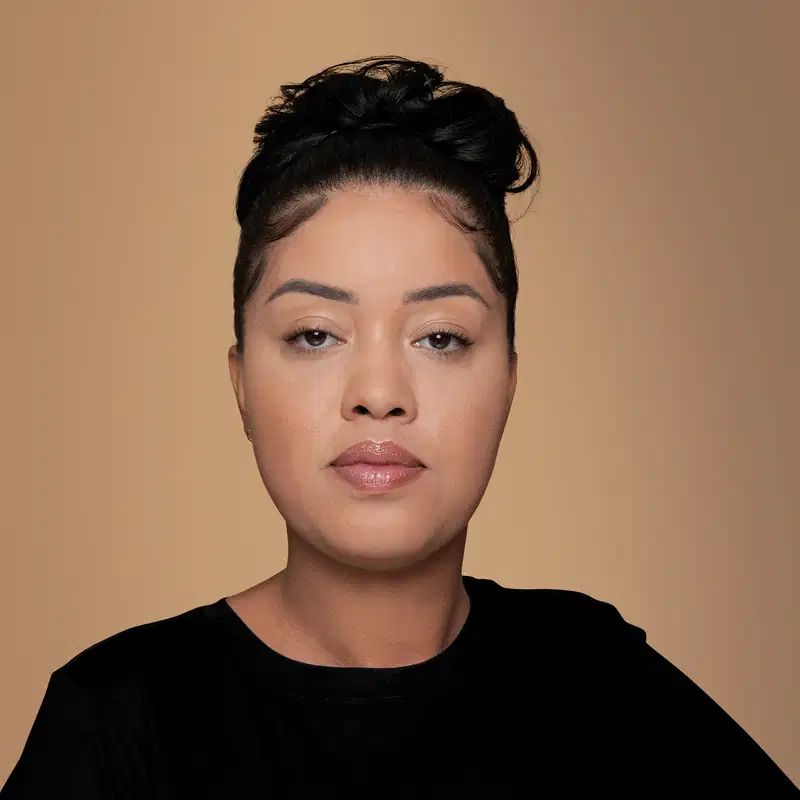 Mulher com cabelo preto presos em um coque, usando camiseta preta, olha sério para câmera contra fundo bege neutro. Ela usa maquiagem bem feita e a imagem sugere que ela está usando a base líquida na cor 170 da marca Make B. Mate Salicylic.