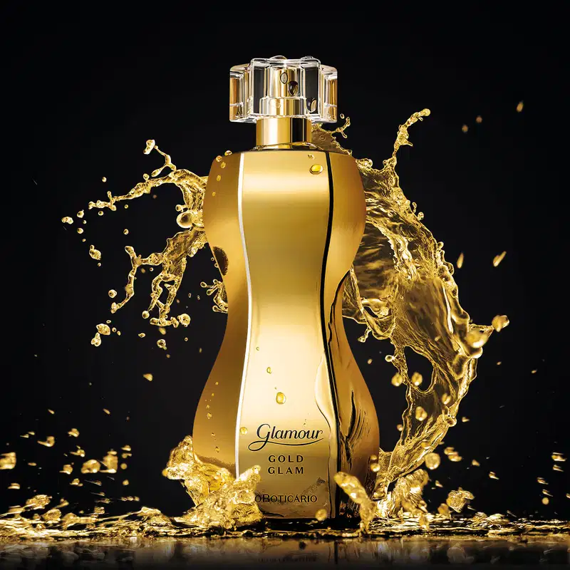 Frasco dourado em formato curvilíneo com borrifador dourado, tampa cristal e letras pretas de desodorante colônia Glamour Gold Glam com respingos de líquido dourado em movimento contra fundo preto.
