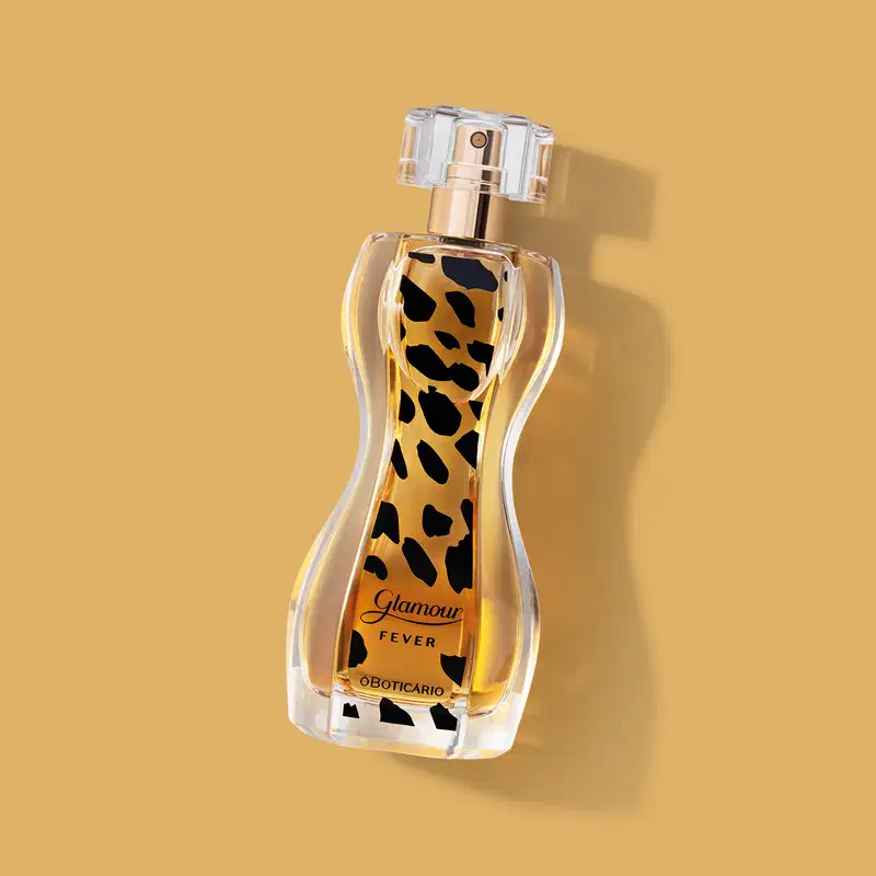 Vidro transparente com formato curvilíneo e líquido âmbar, borrifador dourado, tampa cristal e rótulo com estampa de animal print de desodorante colônia Glamour Fever sobre fundo bege com sombra, spray prateado no topo e líquido âmbar visível.