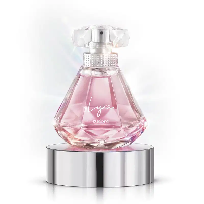 A imagem mostra o Desodorante Colônia Lyra, da Eudora, em frasco de vidro transparente com formato geométrico facetado. O líquido dentro é de cor rosa claro. O frasco repousa sobre uma base cilíndrica de metal prateado.
