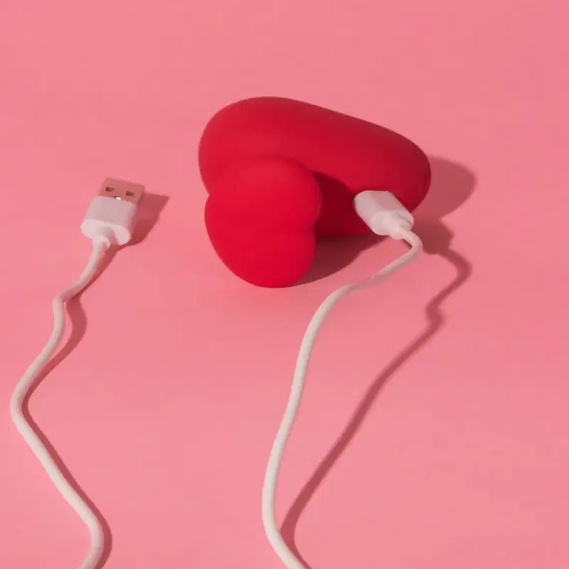 Acessório em formato de coração vermelho com cabo de carregamento USB branco, em fundo rosa sólido. Produto da marca Désir Atelier.
