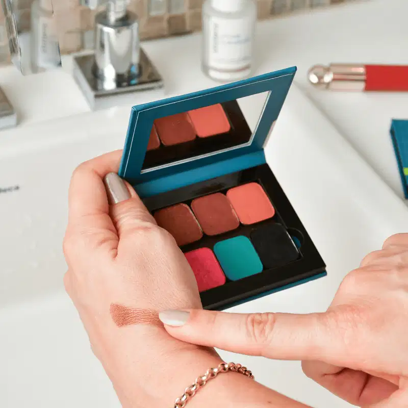 O produto mostrado na imagem é uma paleta de sombras de olhos da marca Statera. A paleta possui um design retangular com uma estrutura em tons de azul escuro. Dentro da paleta, há seis sombras em diferentes tons de vermelho, rosa e turquesa