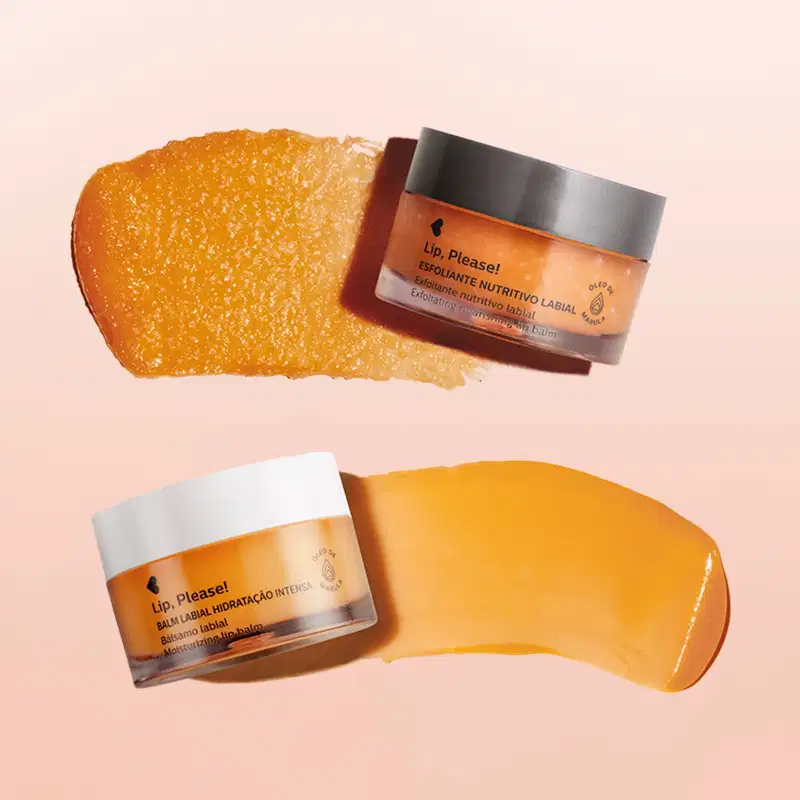Pote de tampa preta de esfoliante nutritivo labial com tampa preta e outro pote de balm labial hidratante com tampa branca, ambos de rótulo laranja e letras pretas com pequena quantidade do produto espalhada ao lado da embalagem em fundo rosa.