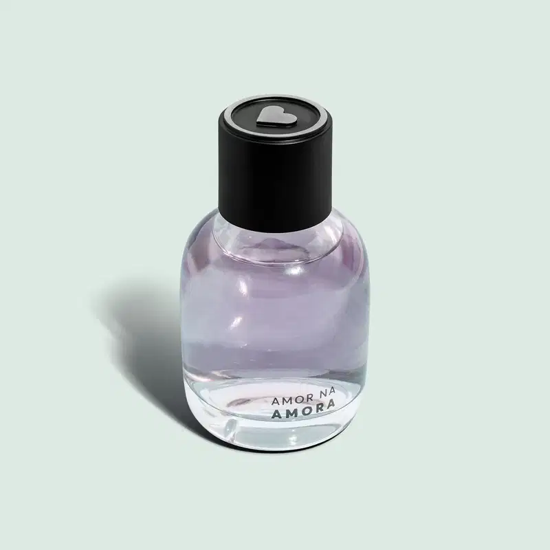 O frasco tem uma tampa preta arredondada. Na parte inferior do frasco, está impresso o texto "AMOR NA AMORA" O produto apresenta uma aparência minimalista, com o frasco transparente permitindo visualizar o líquido lilás em seu interior.